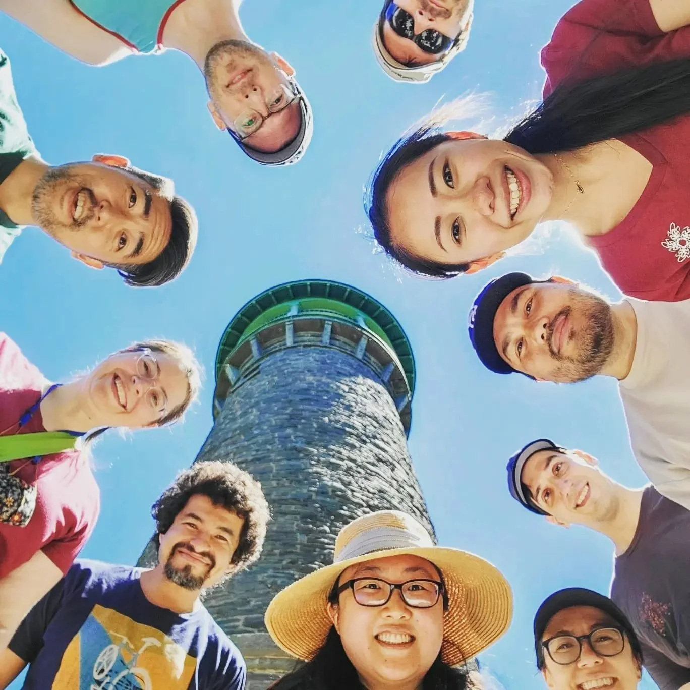 A group of people taking a selfie around a tall stone lighthouse with a green top, looking down at the camera, against a bright blue sky.