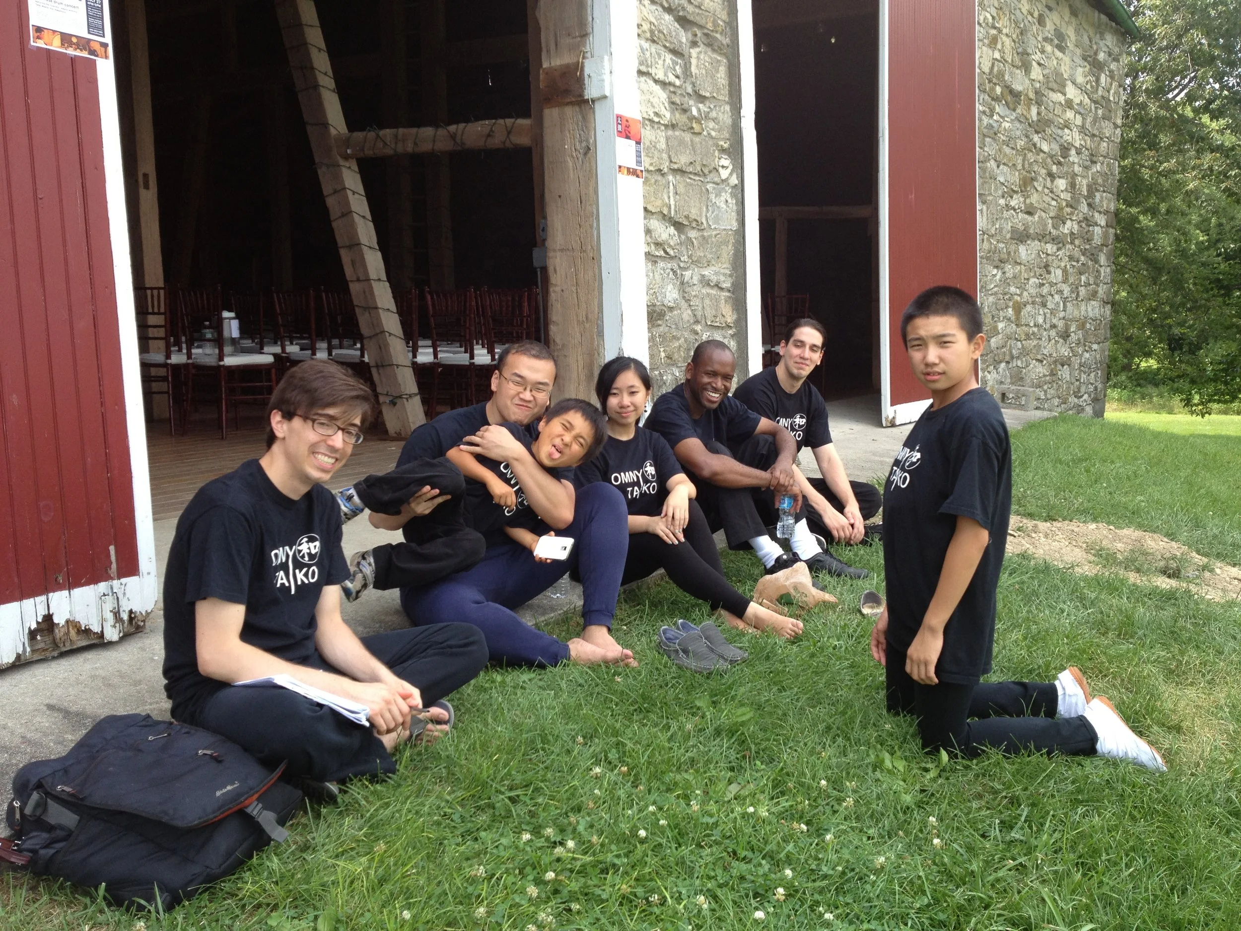 Group of seven people posing outside a barn with an open door, sitting and kneeling on the grass, some smiling and others making playful faces, with chairs and wooden beams visible inside.
