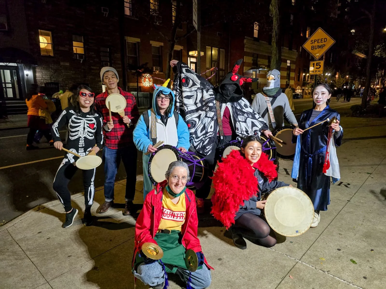 Group of people dressed in Halloween costumes, some with masks and skeleton themes, holding musical instruments such as drums and cymbals, outdoors at night on a city street.