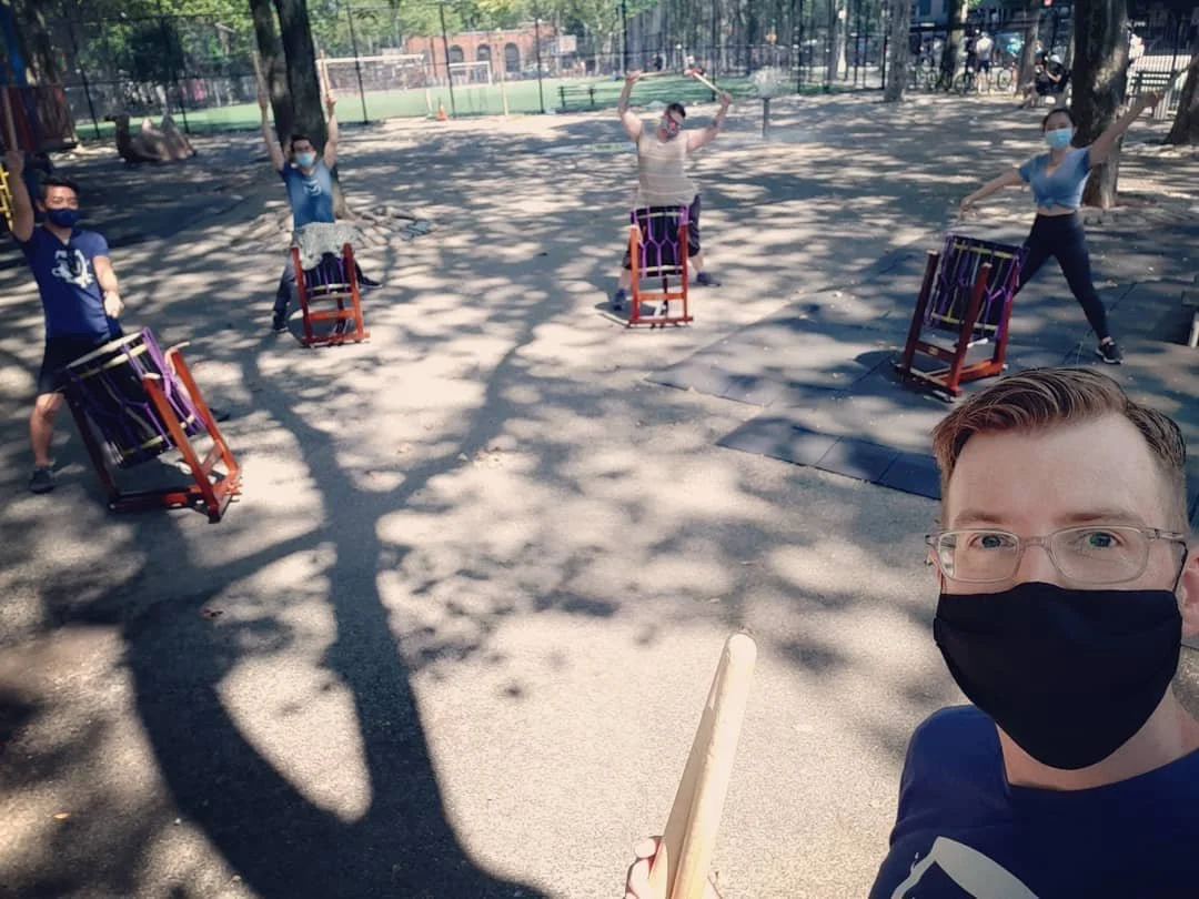 Group of five people practicing drumming on drums outdoors in a park, wearing masks and standing in a semicircle, with one person taking a selfie in the foreground.