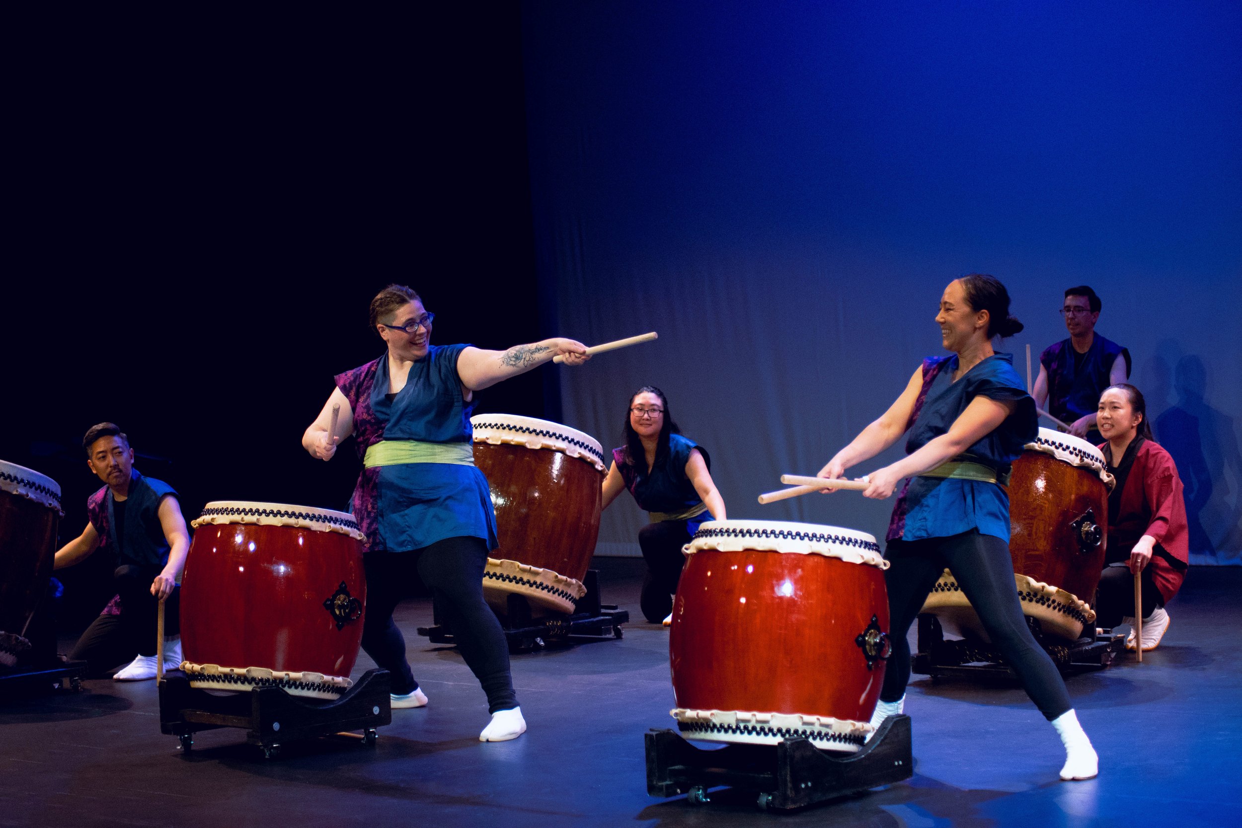 People playing taiko drums on stage, some smiling and engaging in a traditional Japanese drumming performance.