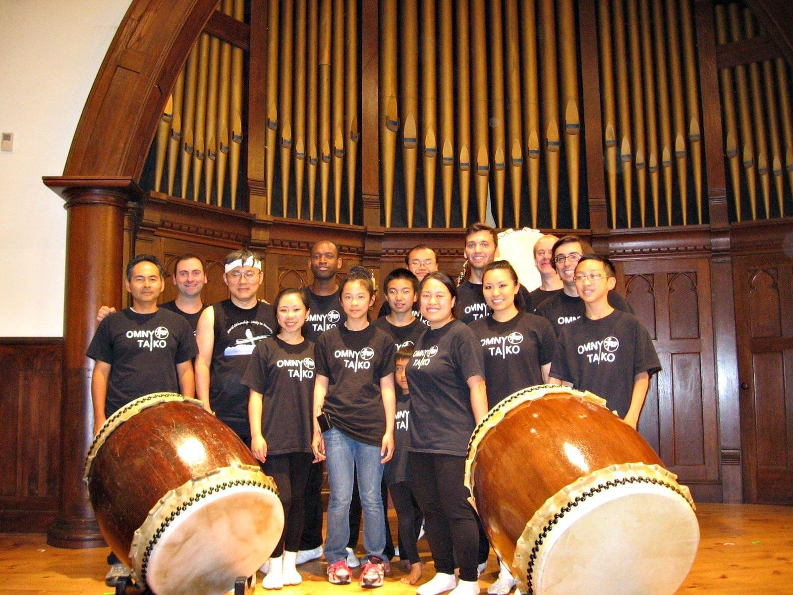 Group of people standing on a stage with large traditional drums and a pipe organ in the background.