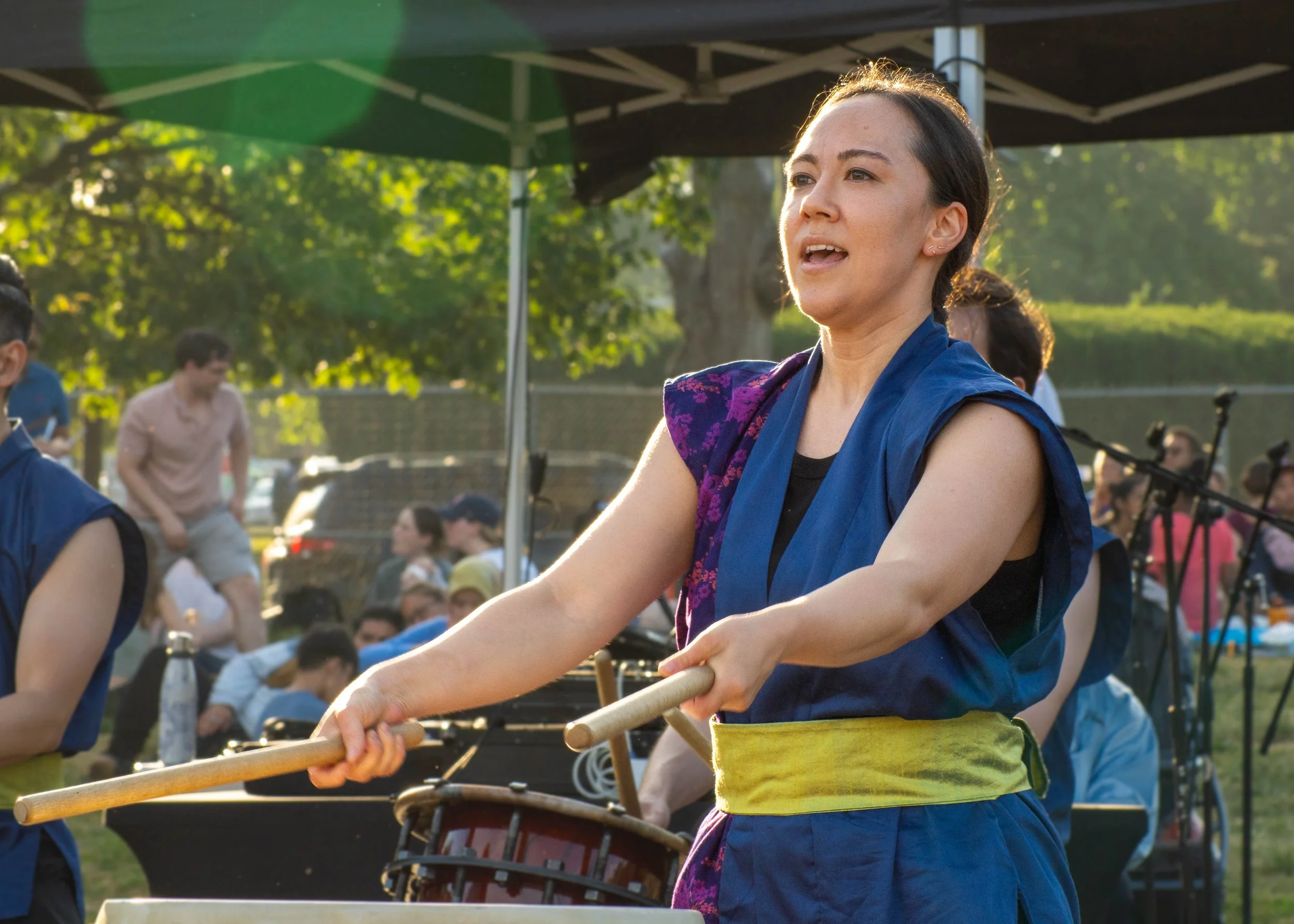 A woman playing a traditional drum during an outdoor cultural event with trees and other people in the background.