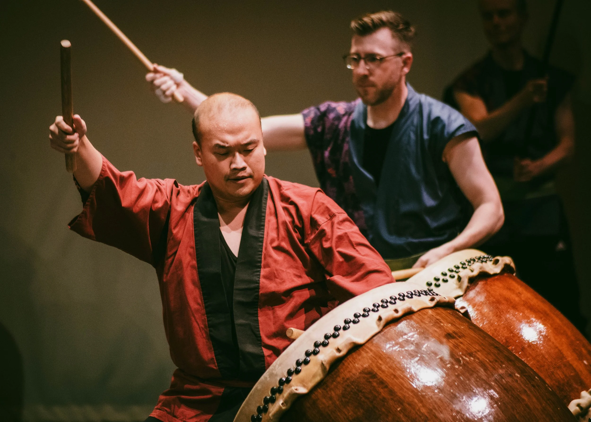 Two performers participating in a traditional Asian drum performance, one in a red and black outfit striking the drum, with a man in glasses and a black shirt with colorful patterns behind him, playing a drumstick.
