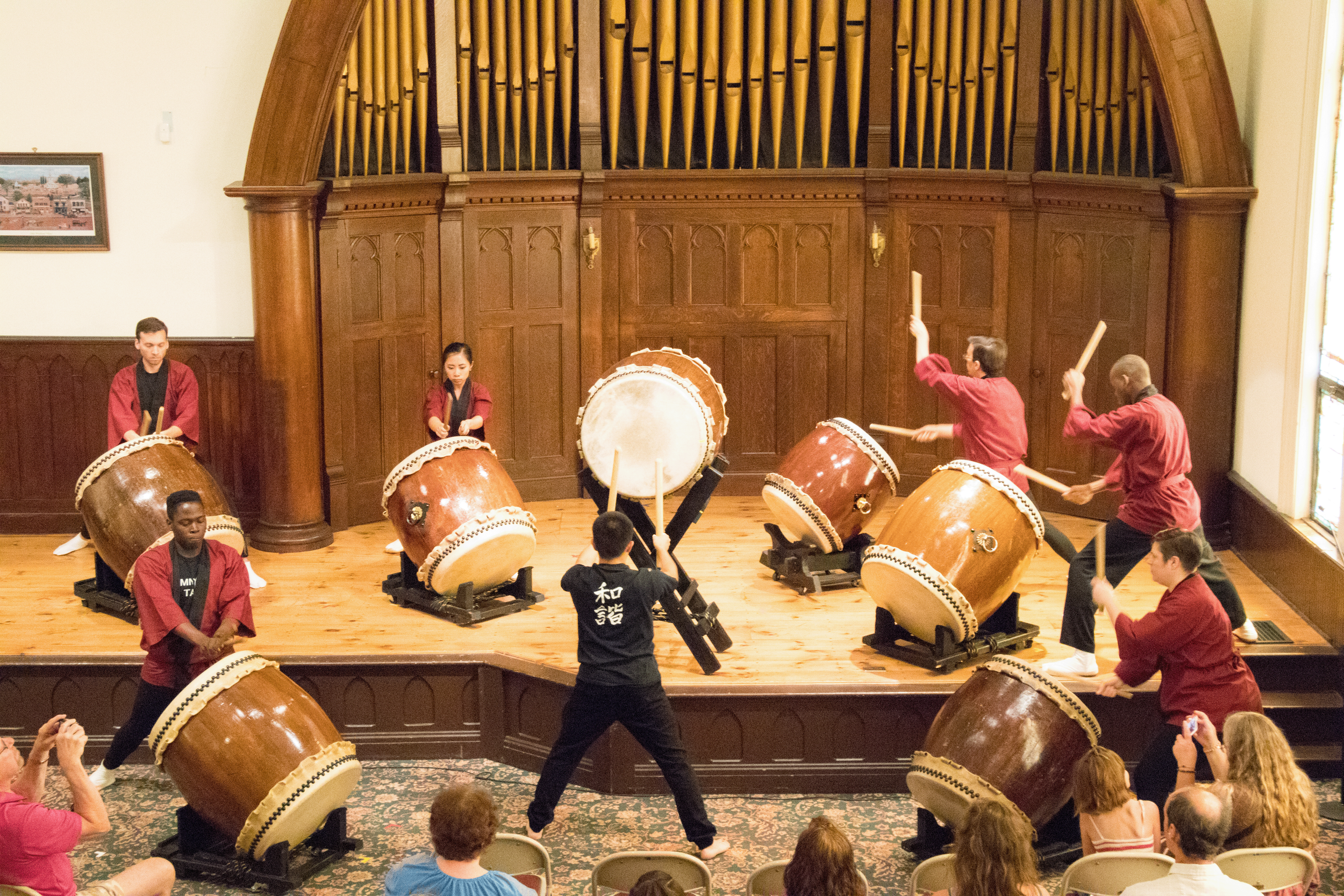 Group of eight people playing traditional Japanese taiko drums on stage in a wooden hall, with an audience seated in front.