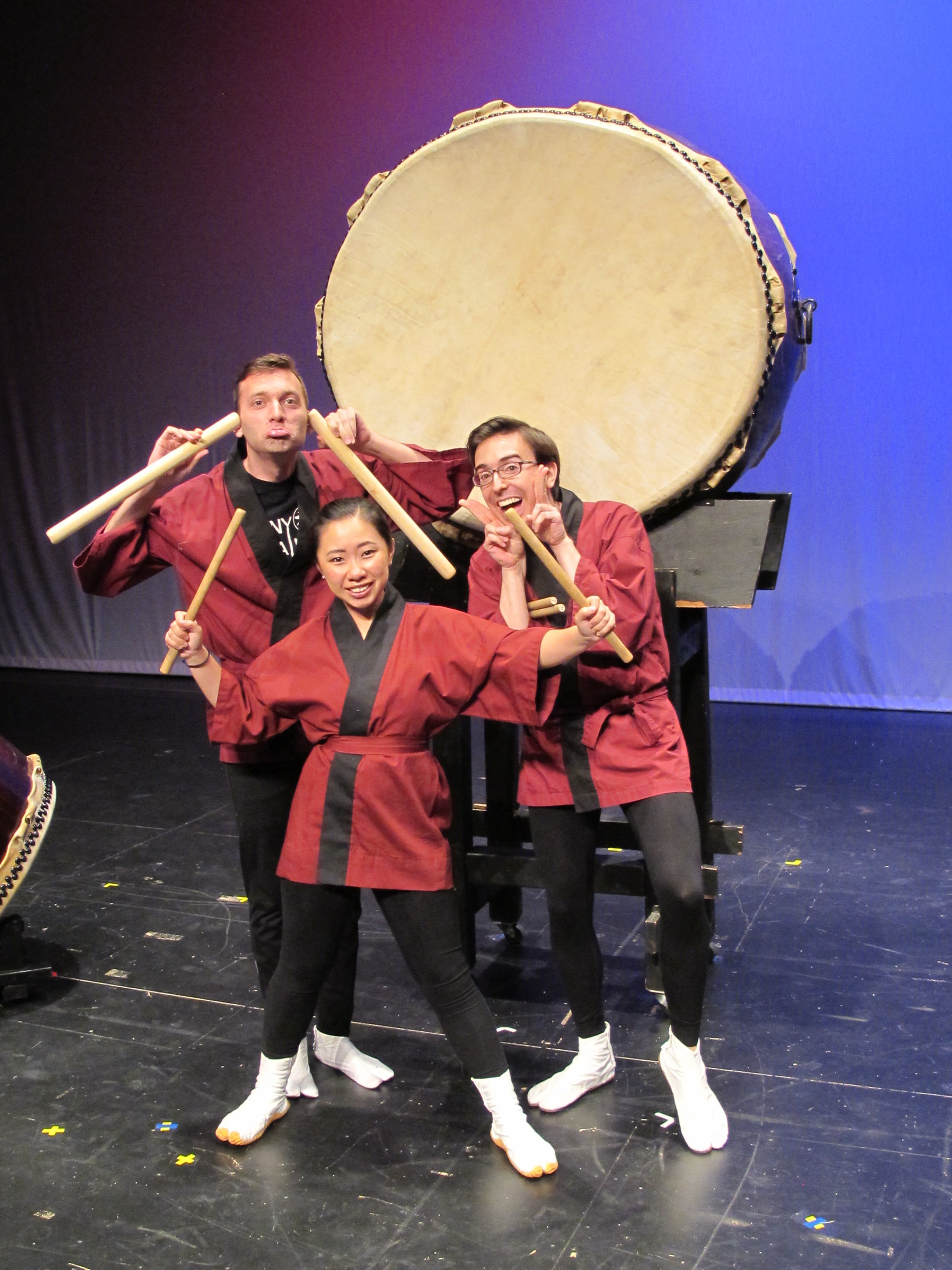 Three performers in red and black costumes posing playfully in front of a large traditional Japanese drum on stage.