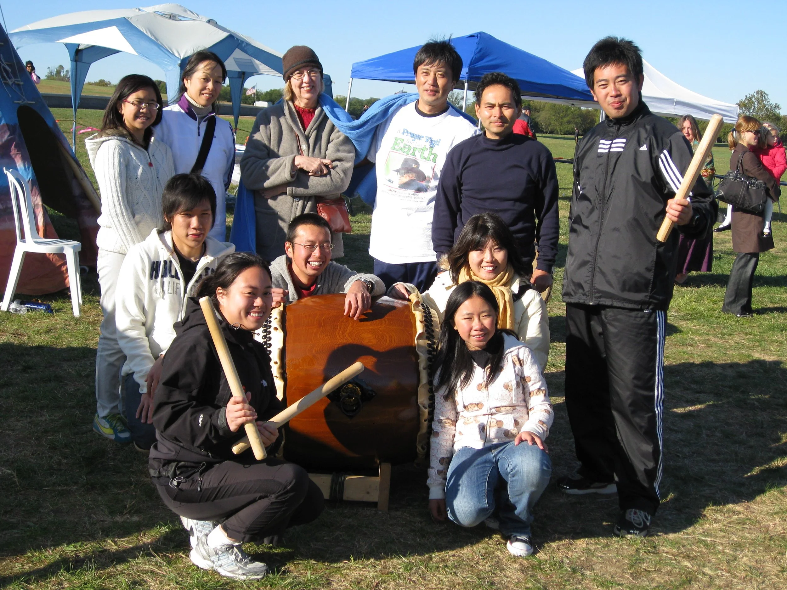 A group of people posing outdoors with a wooden drum, smiling and holding drumsticks, in front of blue and white tents.