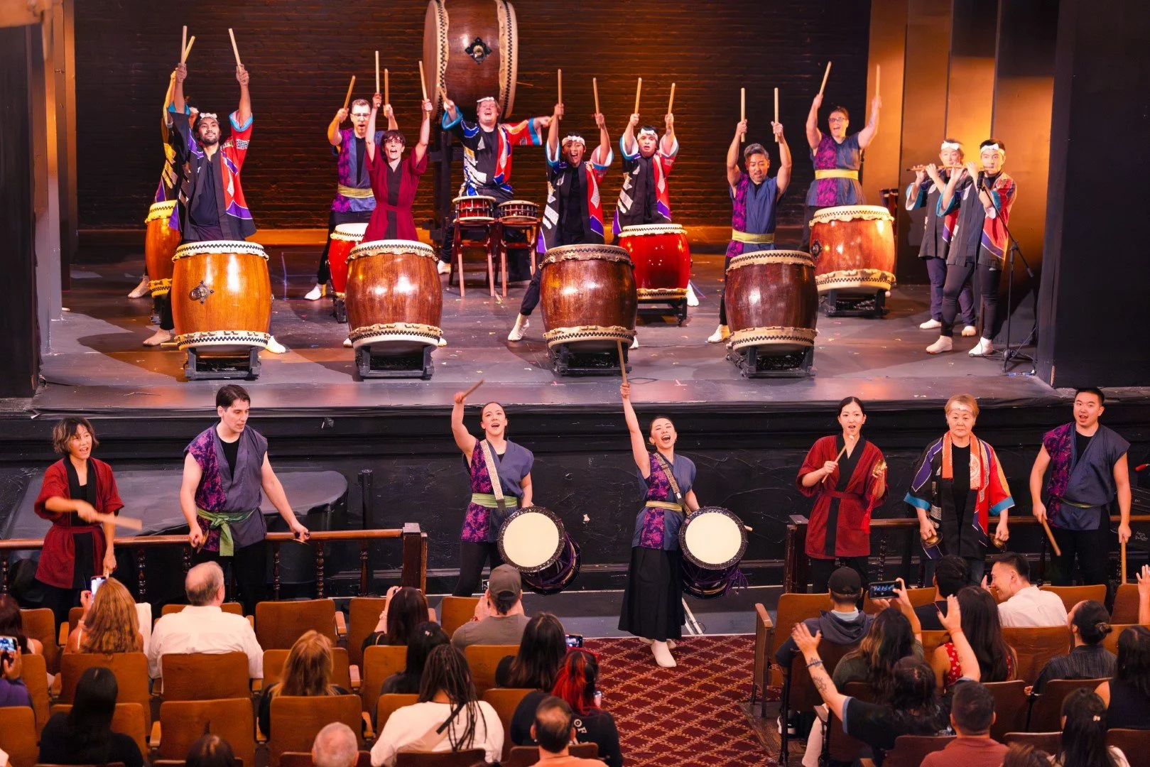 A group of performers on stage playing traditional drums and using sticks, dressed in colorful costumes, with an audience in front watching and taking photos.