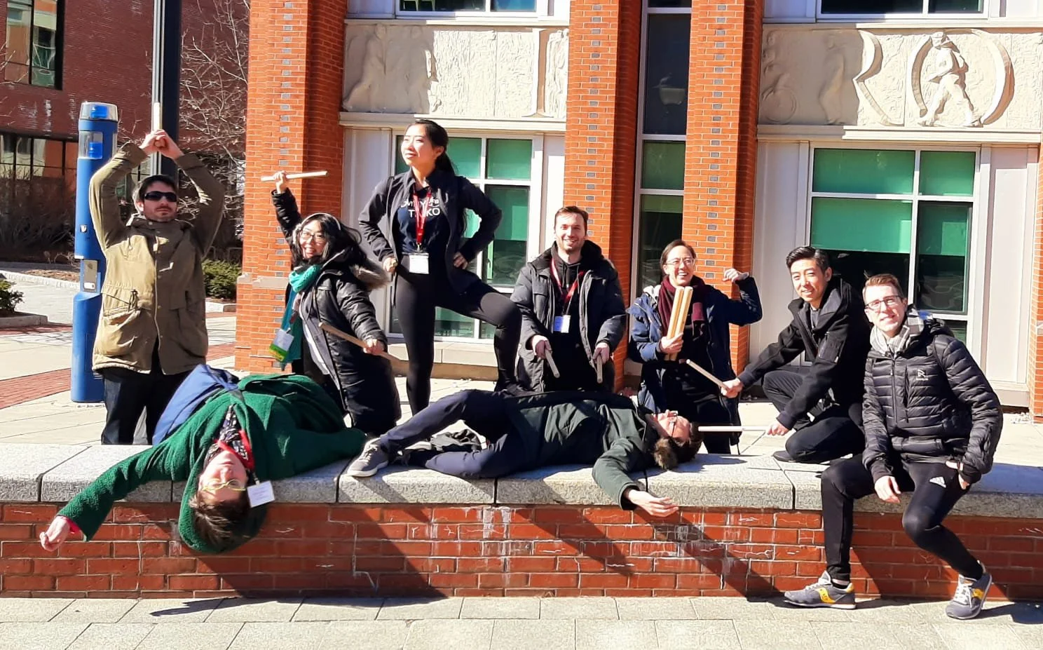 Group of nine young adults posing playfully on a brick ledge outside a building, some lying down and others standing, with some holding sticks or drums, on a sunny day.