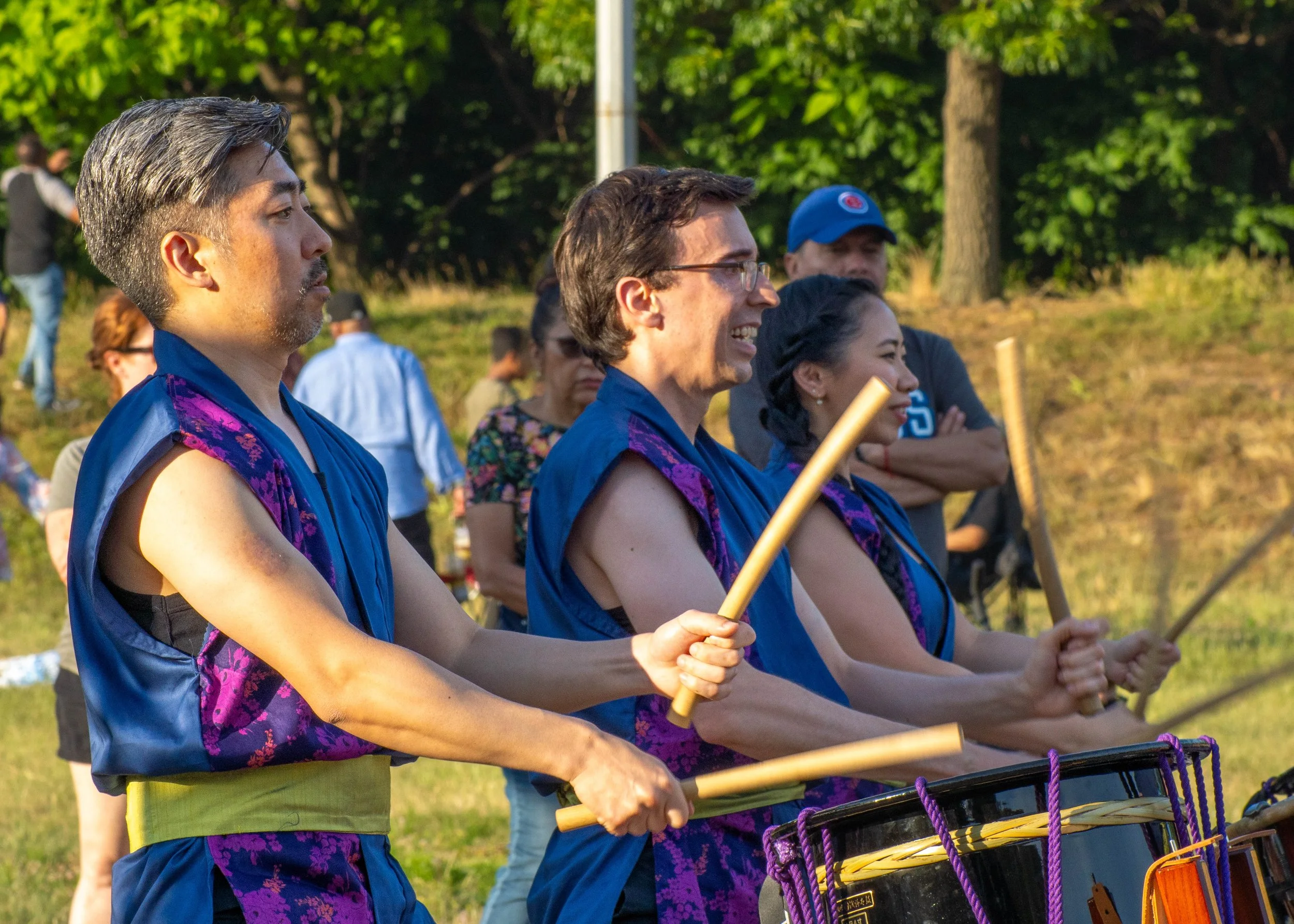 Group of four musicians playing drums outdoors during daylight, surrounded by trees and onlookers.