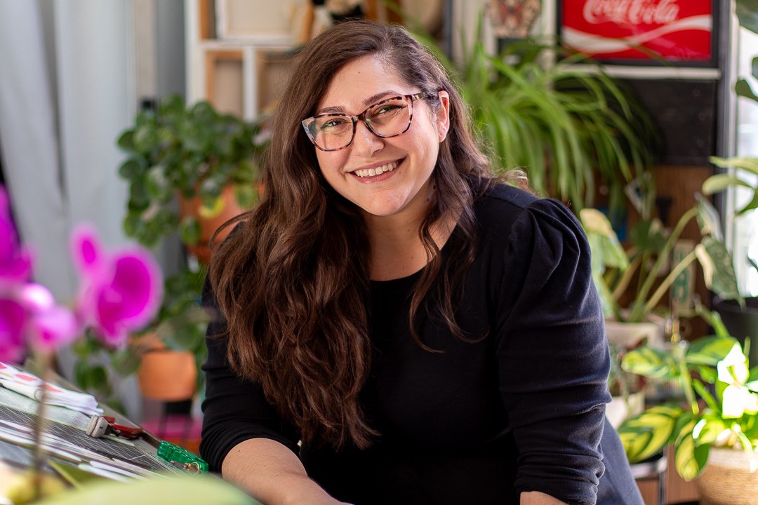A smiling woman with long brown hair, glasses, and a black top, sitting indoors surrounded by green plants and pink flowers.
