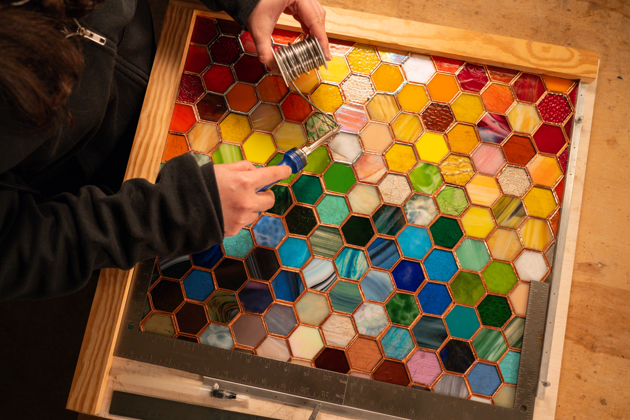 Person creating a stained glass panel with hexagonal glass pieces in various colors, using soldering iron in a wooden frame.
