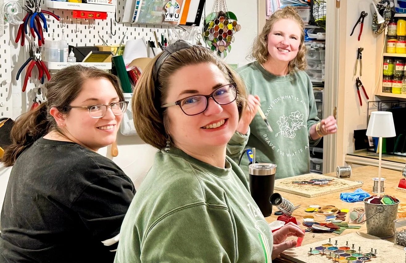 Three women are working at a crafting table in a workshop. They are smiling and looking at the camera.