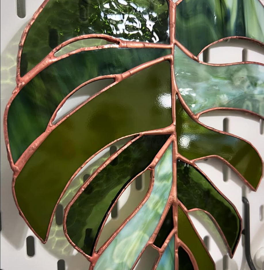 A finished stained glass art piece of a Monstera plant leaf finished in green glass and copper foil edges