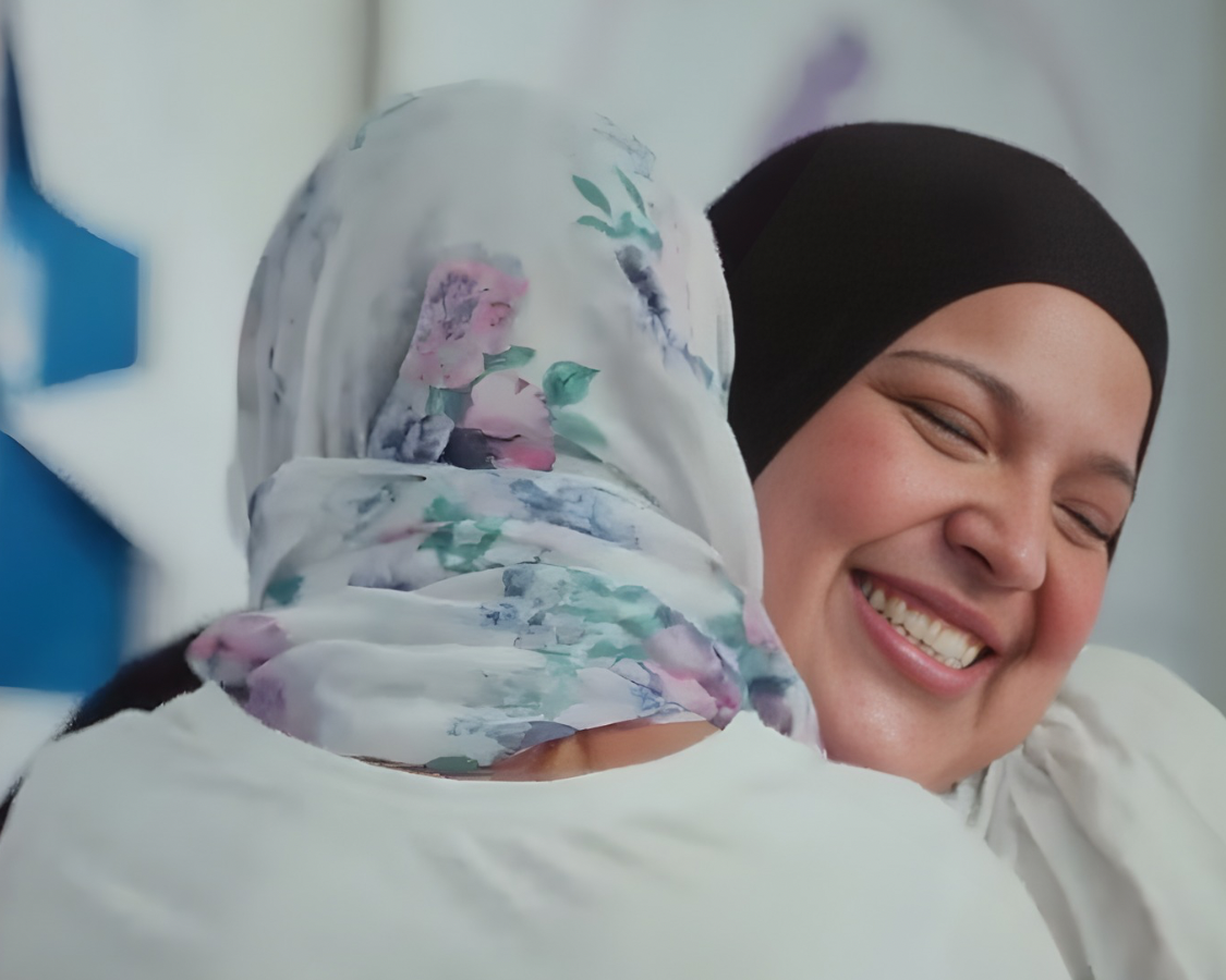 Two women wearing head scarfs hugging each other. One is smiling.  The other one only the back of her head can be seen.  She is the survivor of domestic abuse and anonymous.