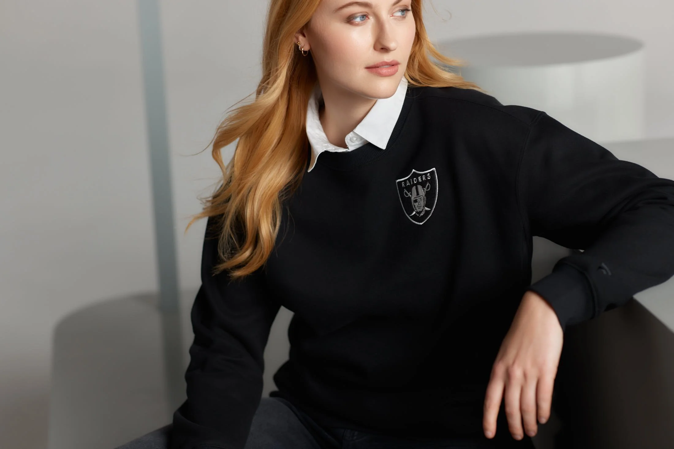 Person wearing a black Raiders sweatshirt with a white collared shirt underneath, sitting indoors.