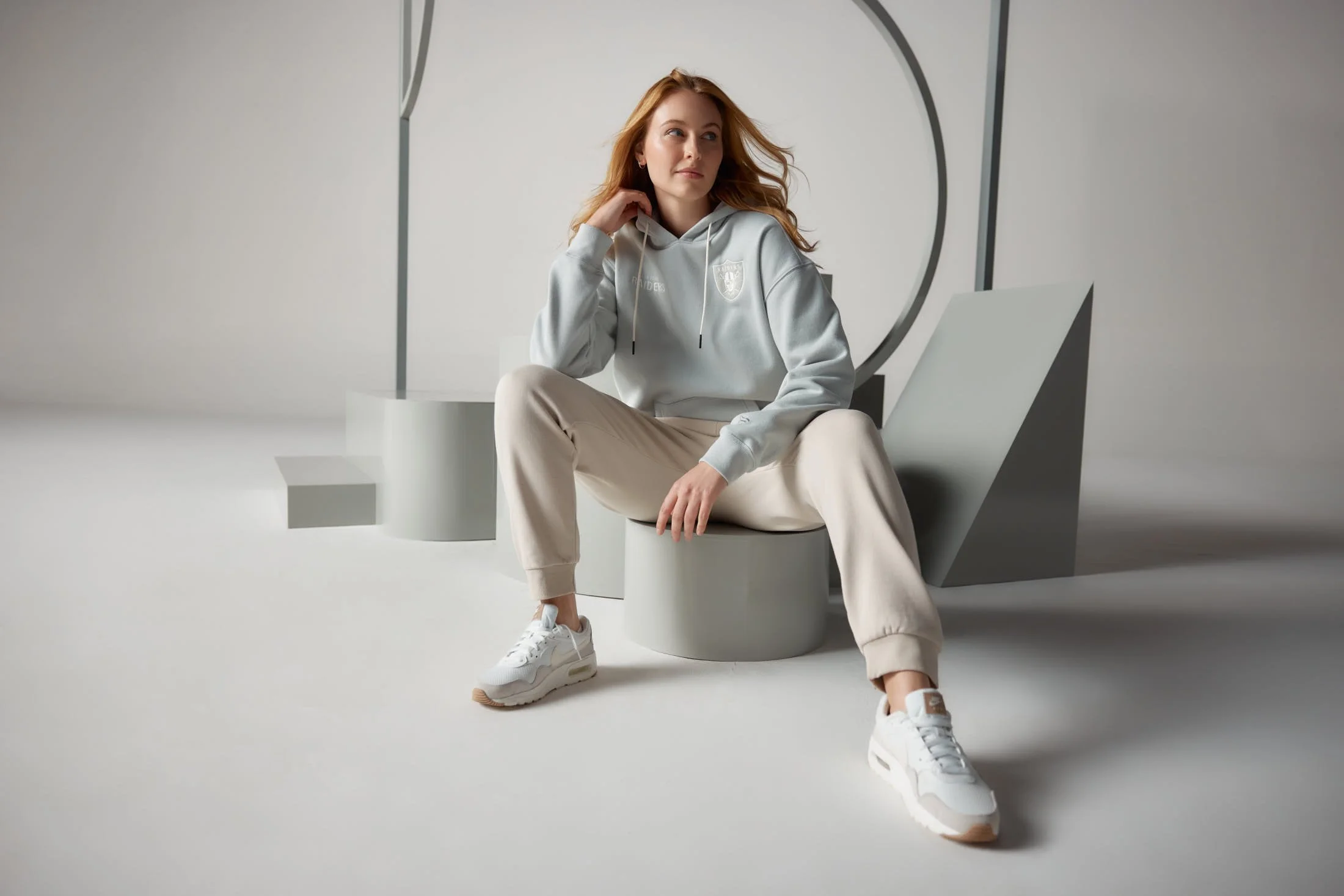 Woman sitting on a geometric stool in a minimalistic studio, wearing a light hoodie, beige sweatpants, and white sneakers.