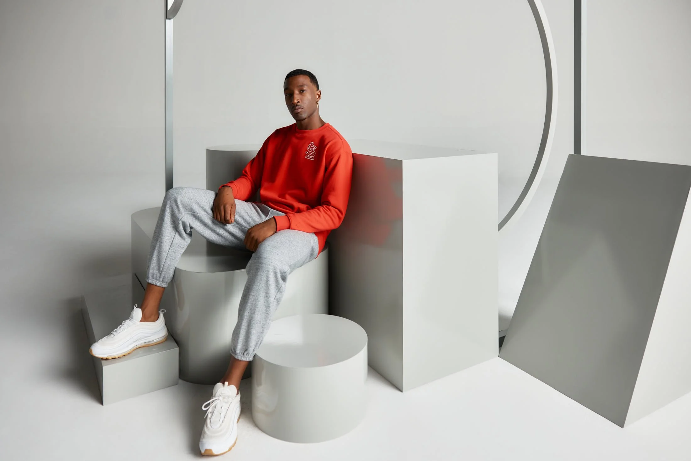 Man wearing a red sweatshirt and gray sweatpants sitting on geometric shapes, with white sneakers on.