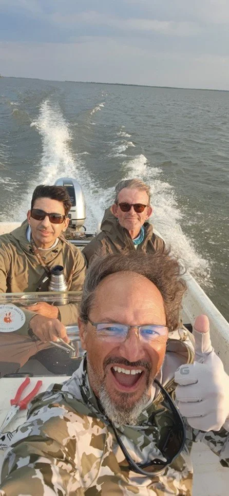 Three men on a boat taking a selfie, with water and boat wake behind them, during daytime.