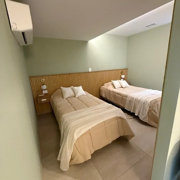 Two beds with beige bedding and pillows in a minimalist hotel room with light green walls, a wooden headboard, wall-mounted bedside lamps, and an air conditioning unit.
