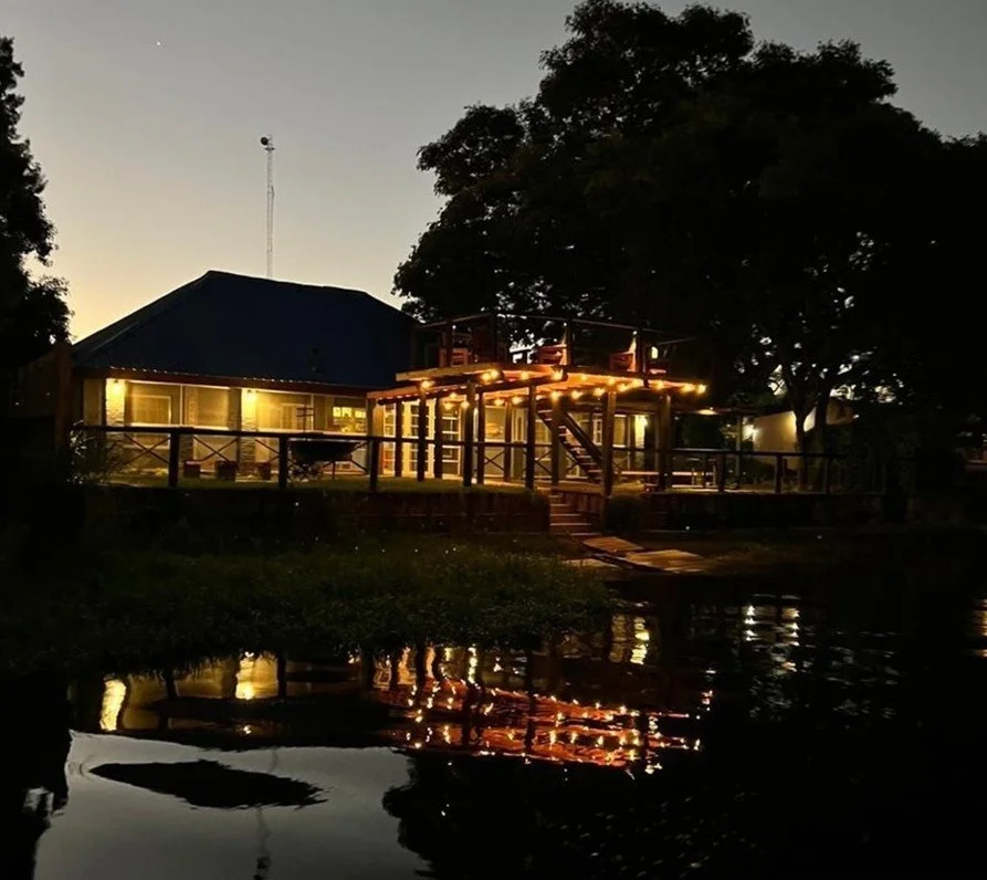 A house at dusk with warm string lights on the porch and deck, reflecting in a body of water in the foreground, surrounded by trees.