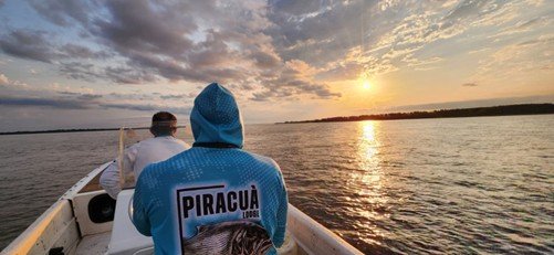 Two fishermen on a boat in a river at sunset, with one wearing a blue hoodie that has 'PIRACUÁ' on the back.