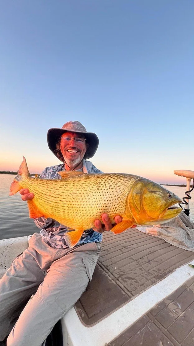 Man in outdoor attire holding a large fish on a boat with water and a sunset sky in the background.