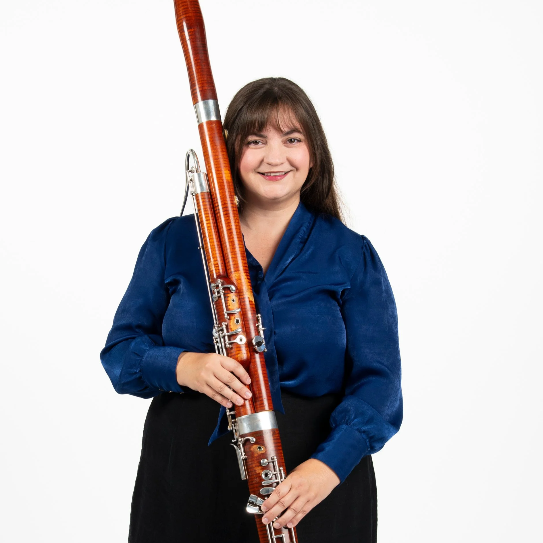Woman with long brown hair wearing a blue blouse and black skirt holding a bassoon.