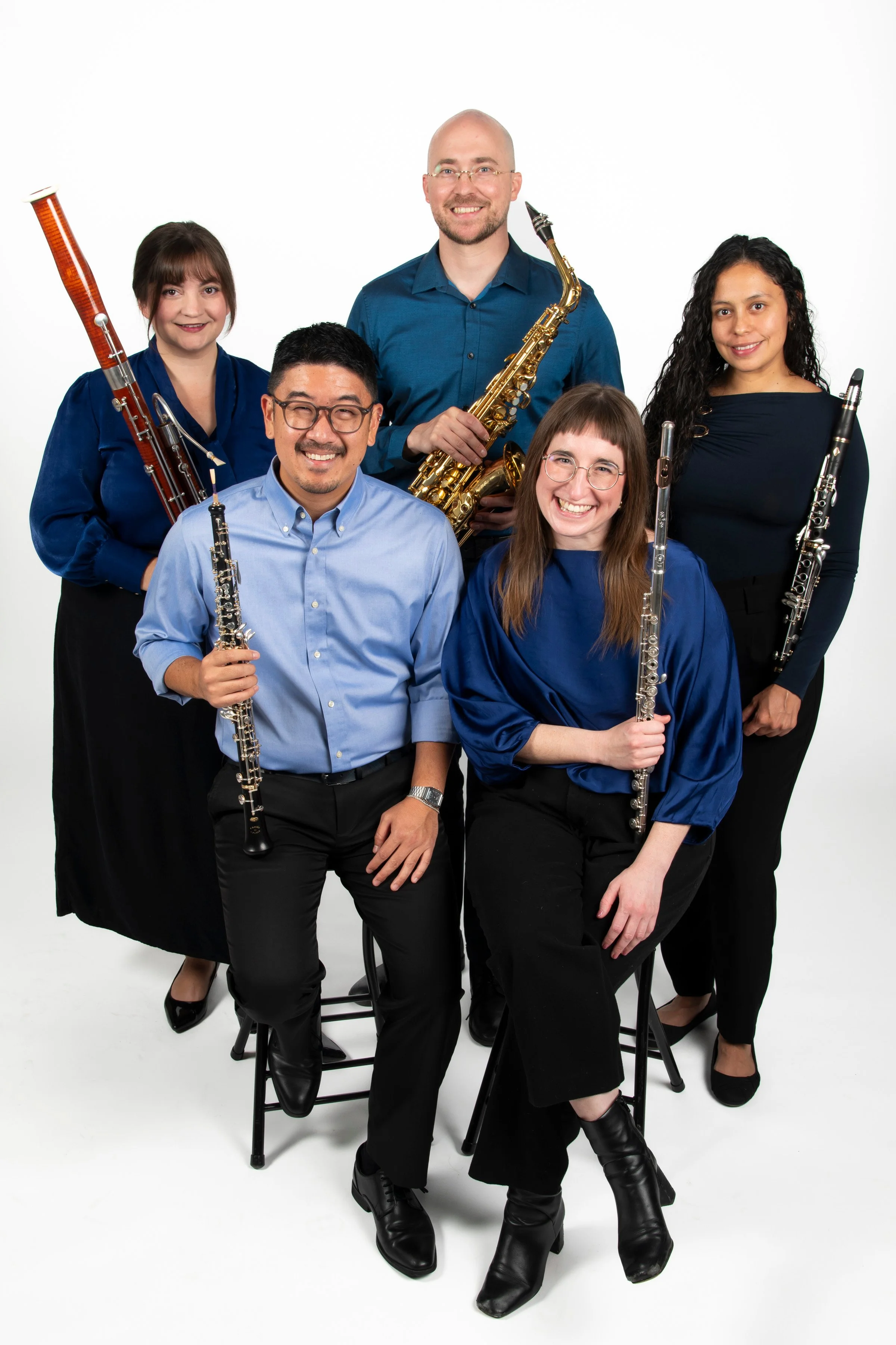 Group of six musicians holding various wind instruments, posing together in a photo studio with a white background.