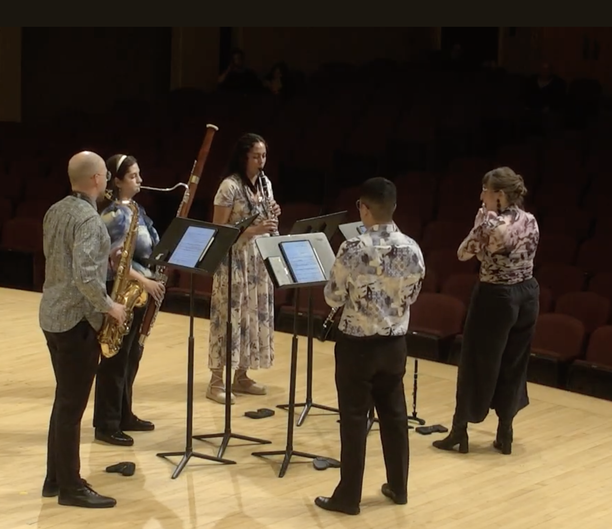 Group of six musicians practicing on stage in a concert hall, with music stands and sheet music, some playing woodwind and brass instruments, and one woman adjusting her hair.