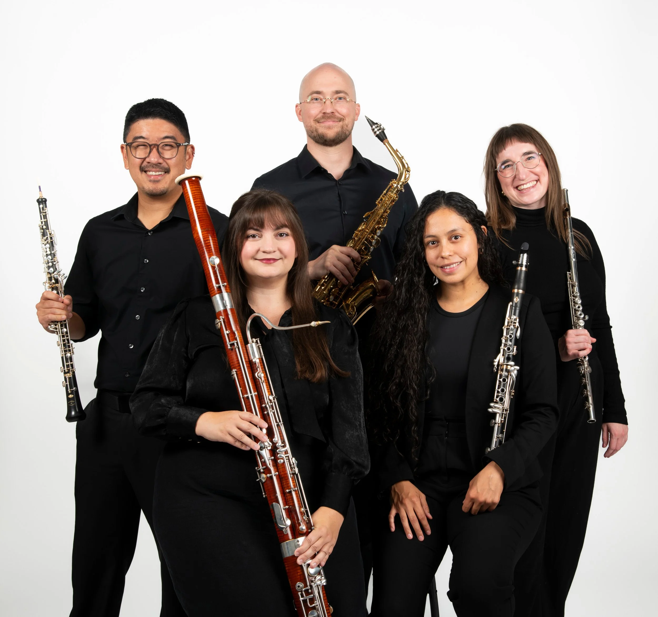 Five musicians holding instruments against a white backdrop, including flutes, clarinets, a bassoon, and a saxophone, dressed in black.
