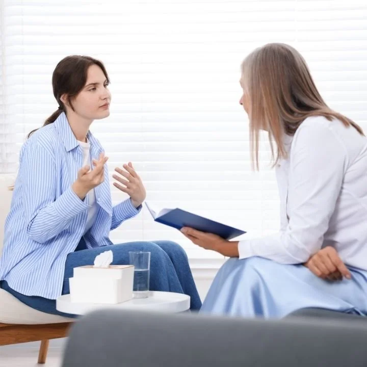 Female therapist and patient seated in conversation during a therapy session with a notebook and tissues on a table.