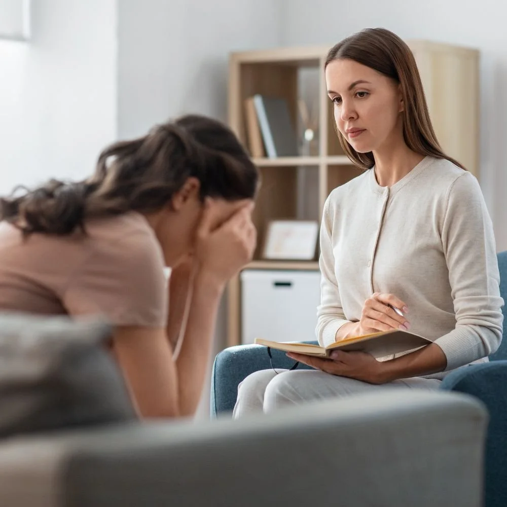 Female therapist holding a notebook attentively listening to a distressed woman sitting with her head in her hands