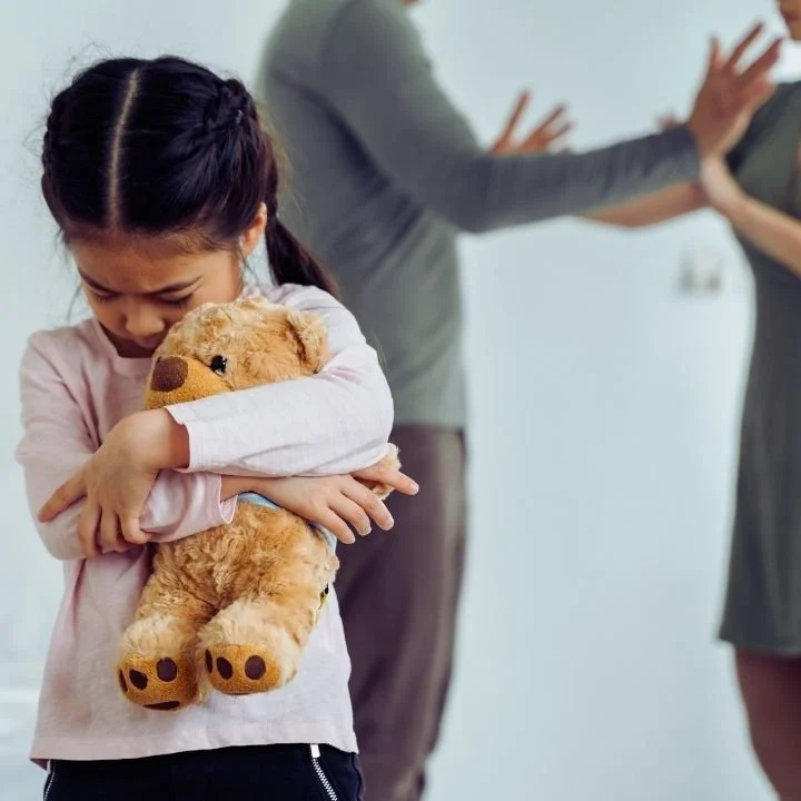 Child holding a teddy bear with head lowered while adults argue in the background.