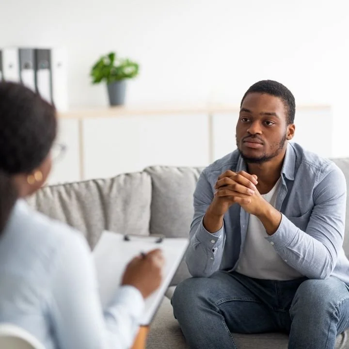 Man seated on a couch facing a therapist who is taking notes on a clipboard.