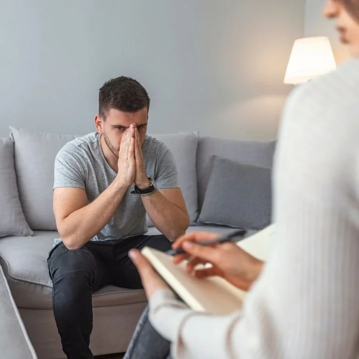 Man seated on couch with hands pressed together near face while therapist listens and takes notes.