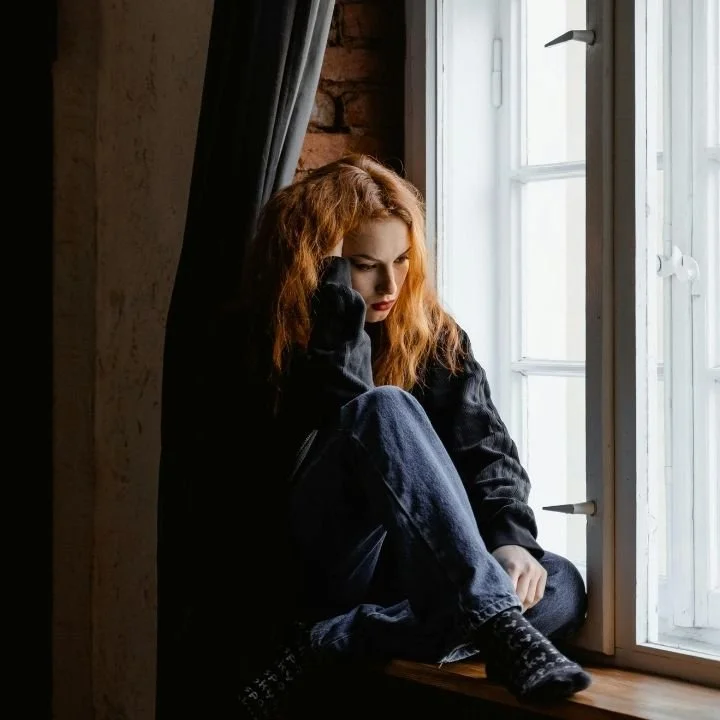 Woman sitting by a window looking sad and hopeless.