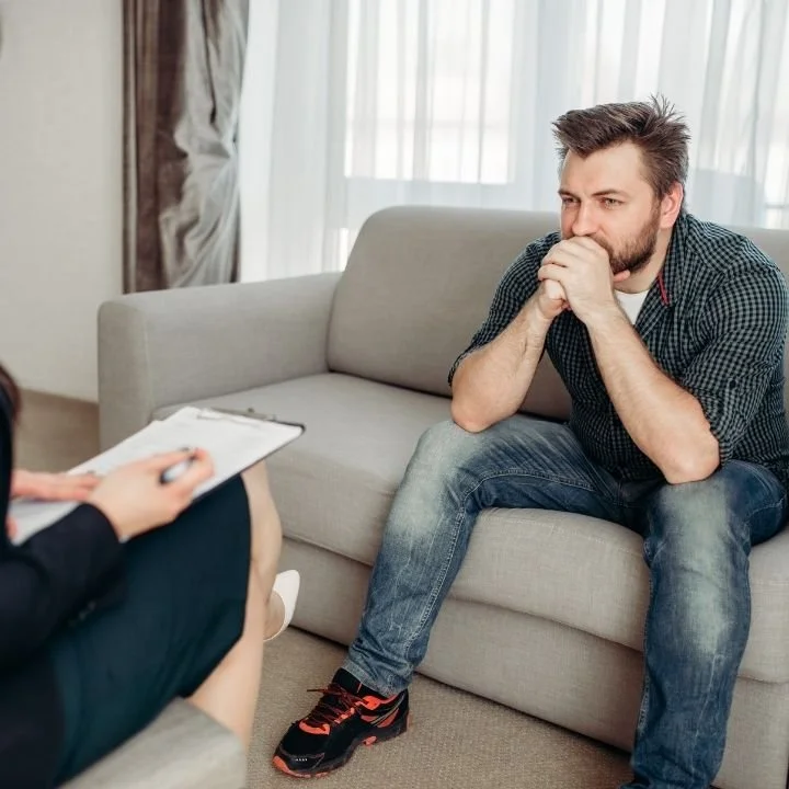 Man seated on couch during therapy session with hands clasped near face, and appearing tense.