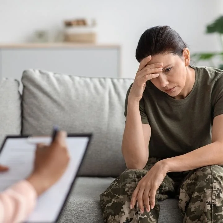 Woman seated on couch with hand on forehead while therapist takes notes nearby.