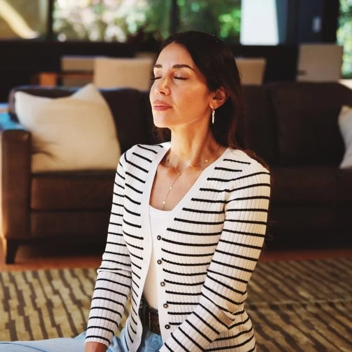 Woman sitting calmly with eyes closed in a living room, practicing mindful breathing.