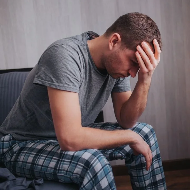 Man sitting on the edge of a bed with his hand on his forehead.