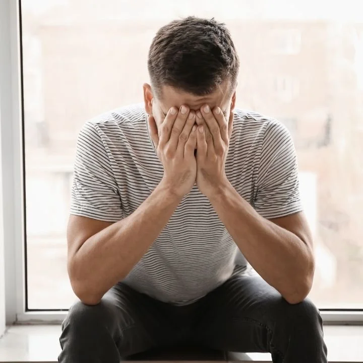 Man sitting by a window with his face covered by his hands, appearing overwhelmed.