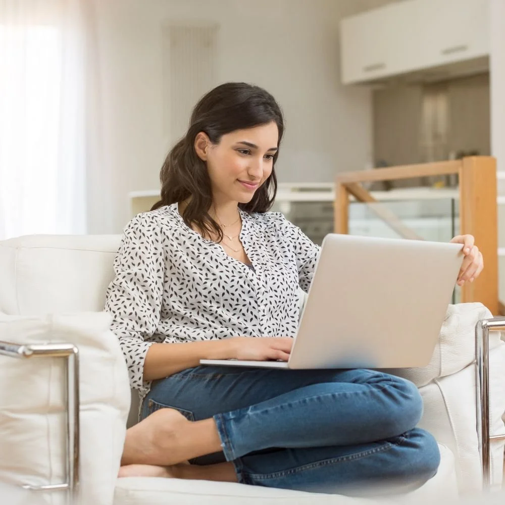 Young woman sitting cross-legged on a white sofa smiling while using a laptop
