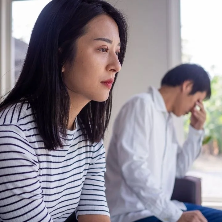 Woman sitting in foreground with distant expression while partner sits behind his holding head.