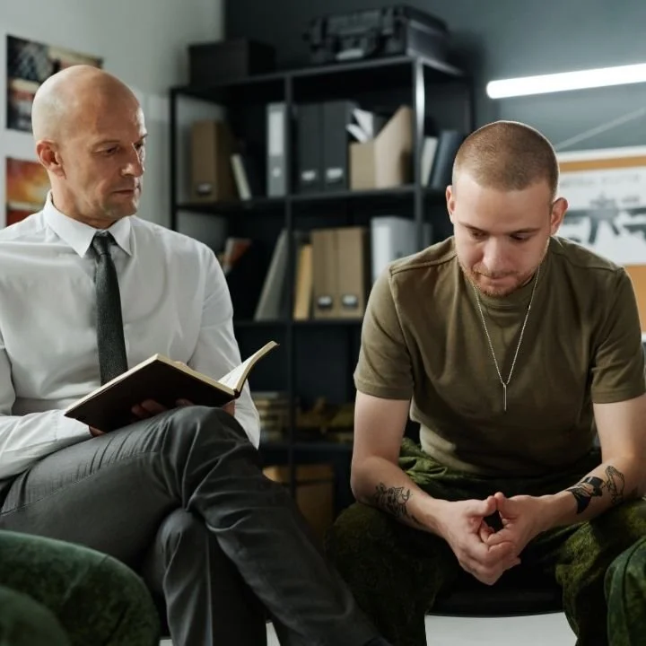 Therapist holding notebook listening to man in military clothing during therapy session