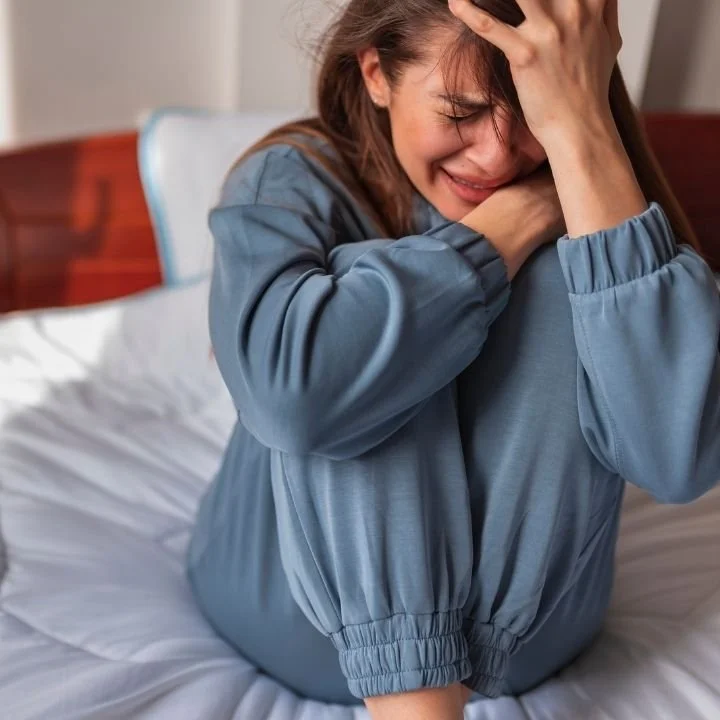 Woman sitting on bed with arms wrapped around herself and crying.