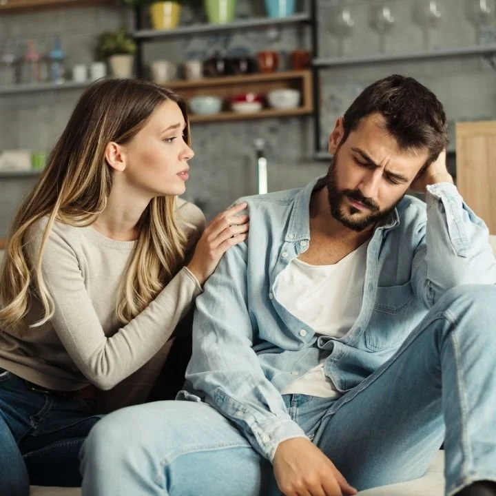 Woman placing hand on partner’s shoulder while he sits with tense, withdrawn posture.