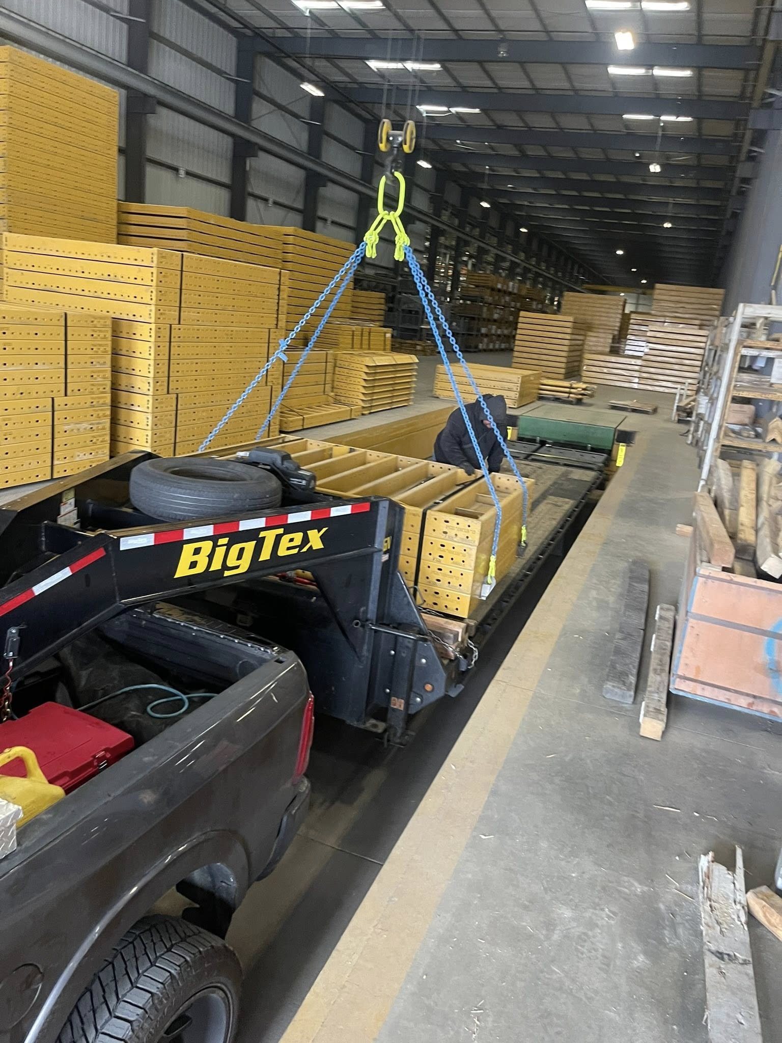 Inside a warehouse with stacks of yellow wooden boards and a trolley with chains hanging from the ceiling, a black vehicle is parked nearby with tools and equipment in its bed.