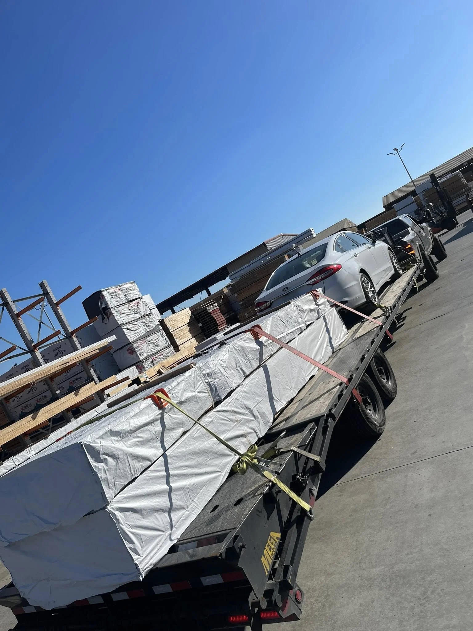 A flatbed trailer carrying large wrapped building supplies, including lumber and construction materials, parked in an outdoor lot with a building and cars nearby under a clear blue sky.