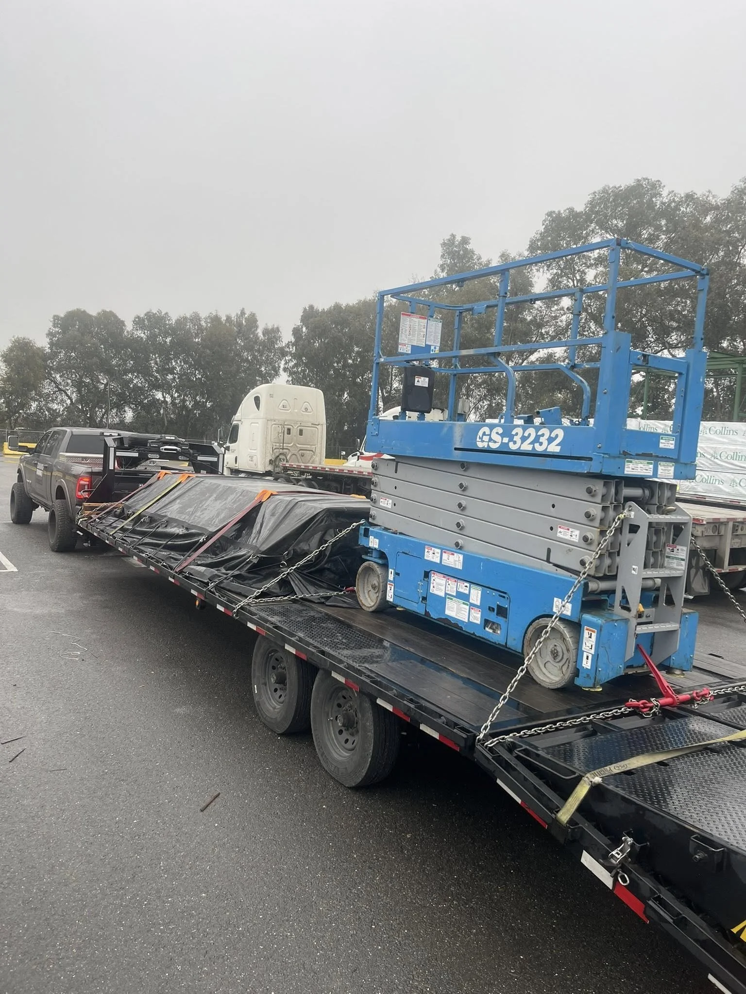 A flatbed trailer carrying a blue scissor lift and a white semi-truck cab in a parking lot, with trees and fog in the background.